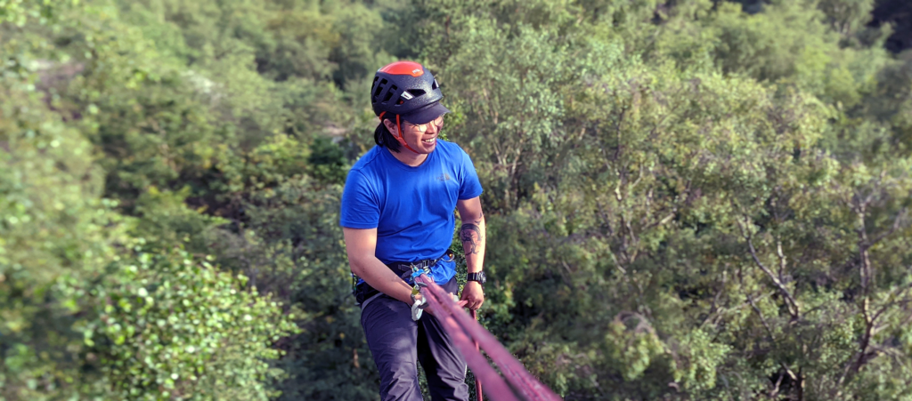 Morning Brief - we'll meet at Basecamp at 08:30 for weather brief and plan the day as a group. Consolidating Group Rigging Skills -varied practice and a look at problem solving and prevention on outdoor crags. Group Abseils - rigging and running group abseils and the common pitfalls to avoid with regards problem solving. Return to Basecamp - return to Basecamp for around 16:00, hand in kit, grab a well-earned brew and review the day. What Next? - your instructor will discuss a personal Action plan and we'll email you some post course resources to help you on your Rock Climbing Instructor journey. Rock Climbing Instructor wearing a climbing helmet and blue t shirt abseiling on a climbing rope from a single pitch crag with a forest below.